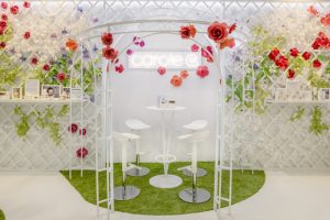 Novembre 2018 – POP-UP STORE – CAROLE G. Dans un environnement joyeux et bienveillant : habillées d’une treille blanche, les cloisons débordent de fleurs en papier, le gazon met en valeur les zones de présentation des produits et une tonnelle métallique vient encadrer le zone d’assise. 